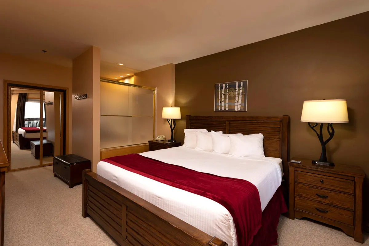 A cozy bedroom centered around a king-sized bed dressed in white linens with a vibrant red throw. Wooden nightstands on either side offer practical storage, while soft pillows and warm lighting suggest an inviting, peaceful atmosphere ideal for unwinding.