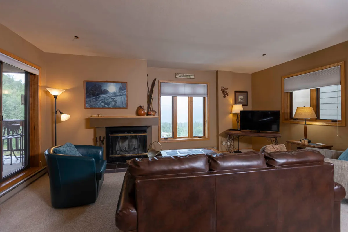 A warm and inviting living room with a wood-burning fireplace as its centerpiece. Plush sofas are arranged around a coffee table, facing large windows that frame scenic views. The room exudes comfort, with rich textures, a cozy rug, and accents perfect for gathering with loved ones. A television is mounted beside the fireplace, completing the entertainment setup.