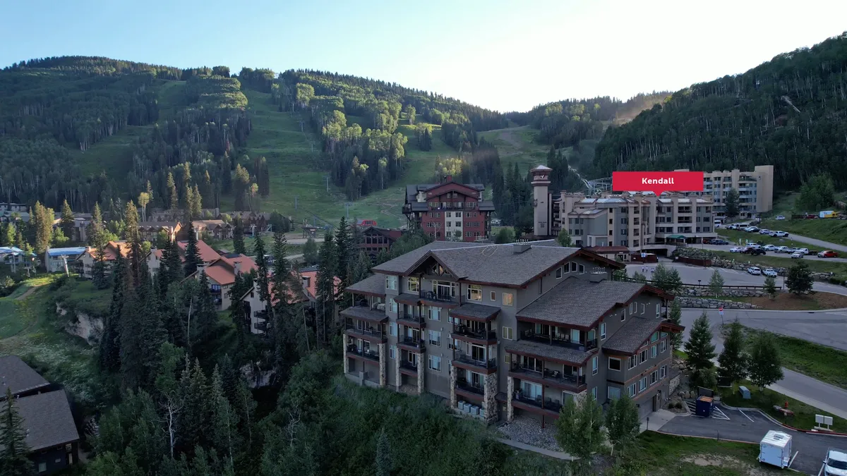 A lively exterior scene featuring residential buildings clustered at the foot of majestic mountains, with a visible parking area and a hint of community activity. The landscape is welcoming, with easy access to mountain adventures and the visual promise of a vibrant village atmosphere.