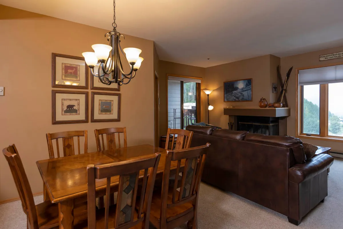 An intimate view of the living area's open layout, featuring a farmhouse-style dining table adjacent to a leather couch and a fireplace. Large windows offer views of the outdoors, while the space is bathed in warm, natural light—ideal for hosting holiday meals, casual dining, or cozy fireside chats.
