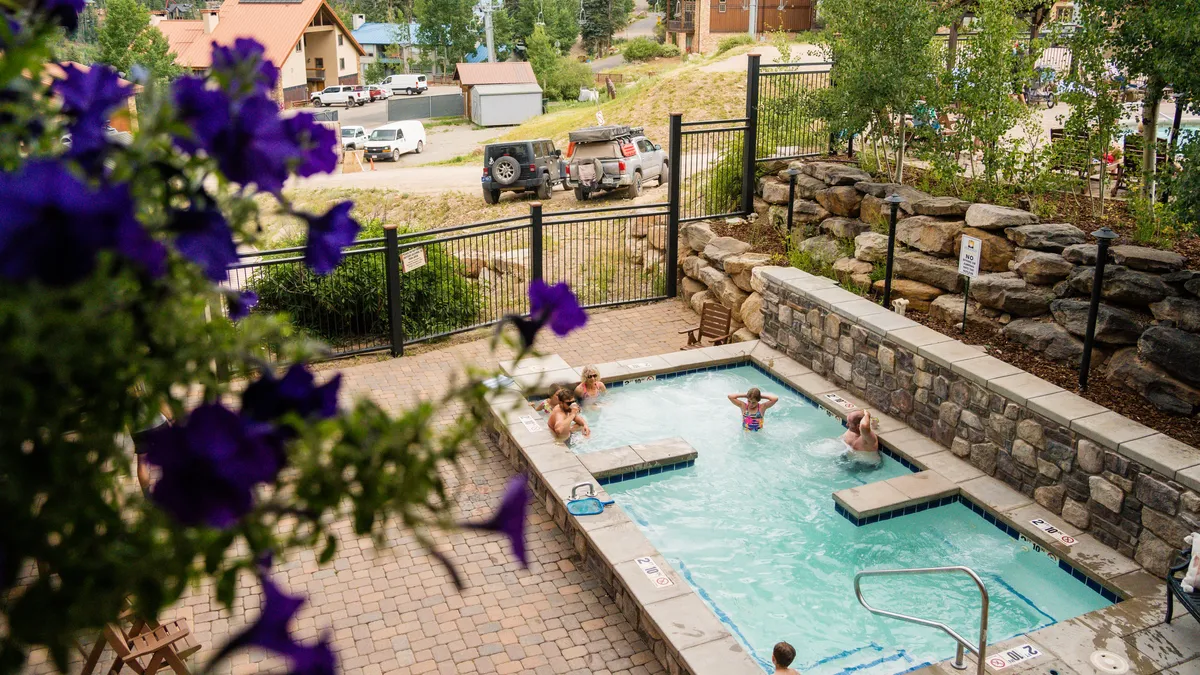 A sparkling outdoor heated pool surrounded by blooming flowers and a natural stone wall. Children and families are joyfully swimming and relaxing, with comfortable seating and lush landscaping enhancing the resort atmosphere.