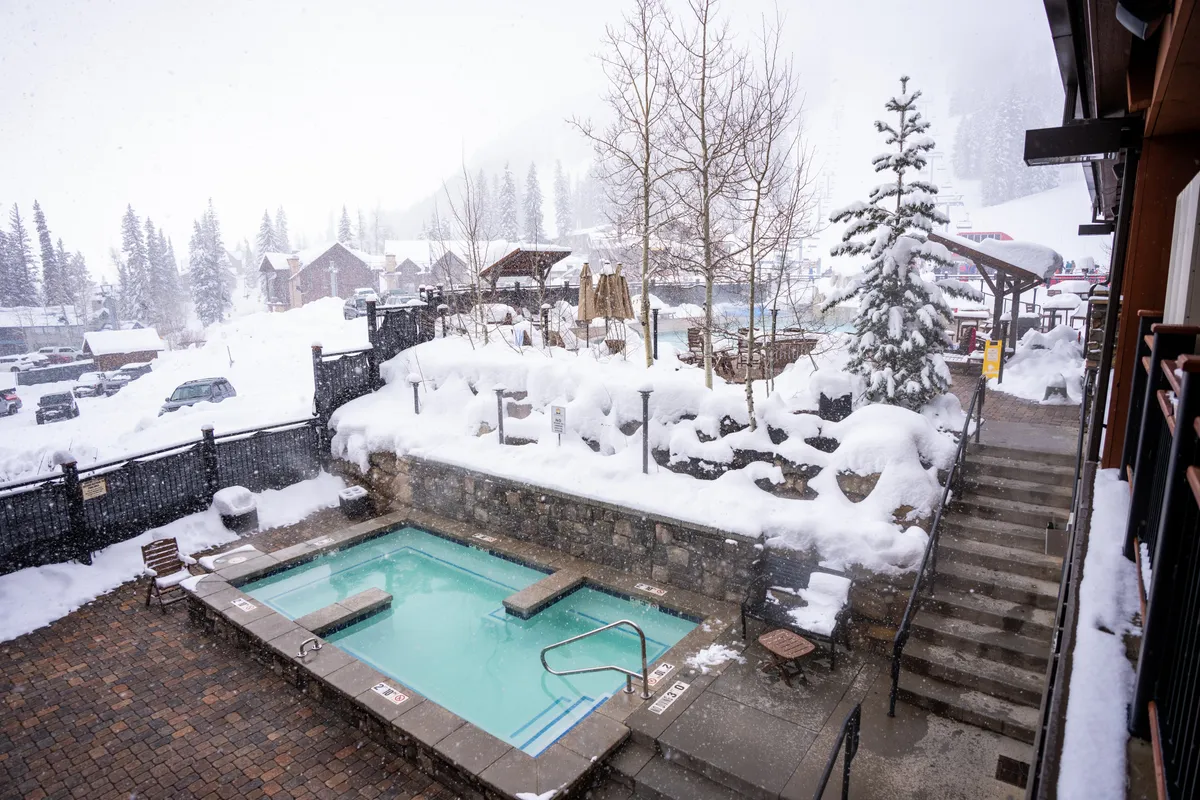 An outdoor hot tub surrounded by a snow-covered landscape and tall trees, with steam rising from the water. The mountains are visible in the background, evoking a sense of relaxation and luxury while immersed in nature.
