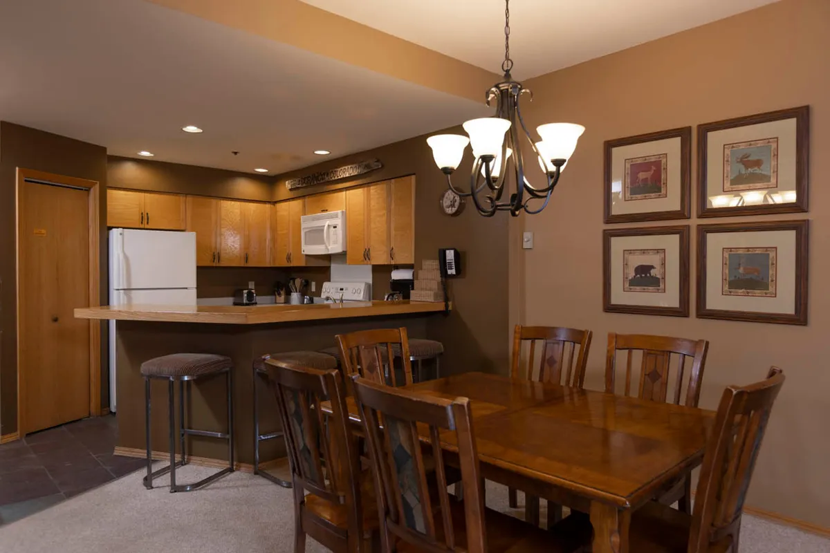 A spacious kitchen and dining area with a prominent wooden table, kitchen counter, and a statement chandelier. The decor feels homey yet elegant, creating a perfect backdrop for shared meals, celebration, and culinary creativity. Framed art adds a personal touch to the walls.