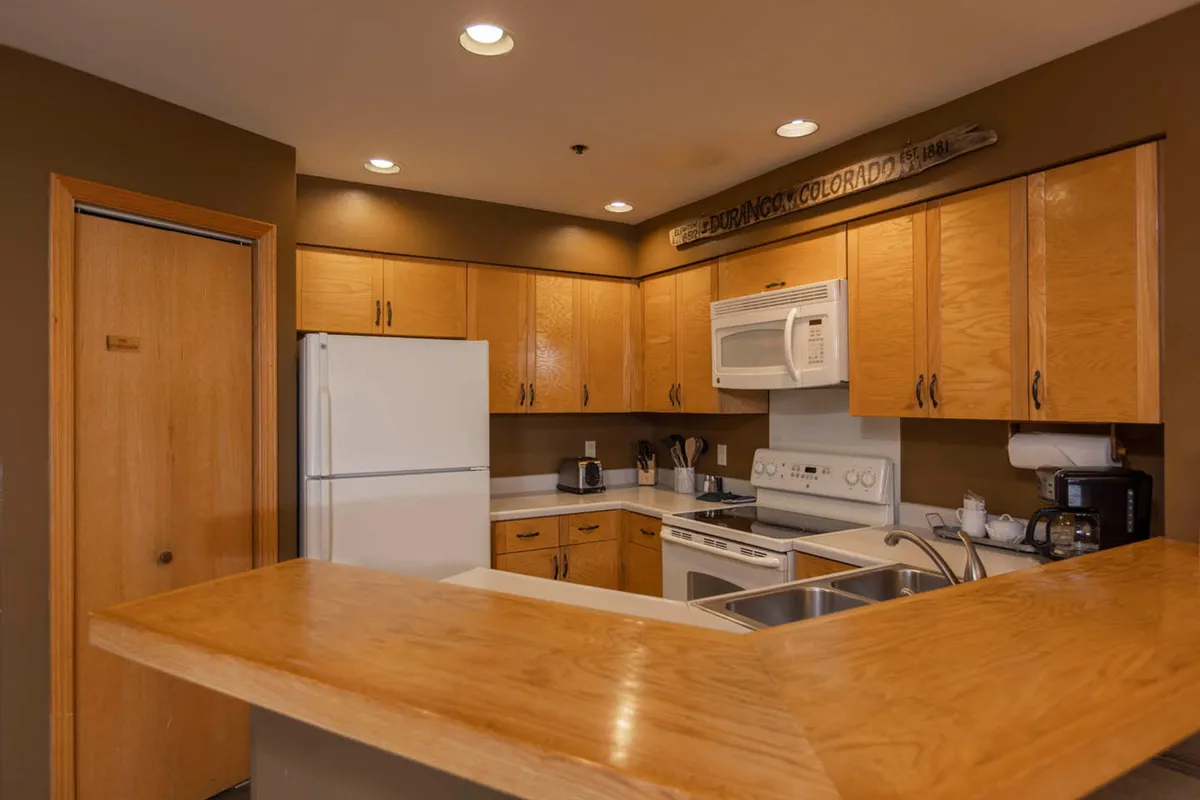 A warmly lit kitchen interior featuring wooden cabinets, white appliances, and a welcoming bar counter. The space is set up for cooking and socializing, with subtle touches that evoke both comfort and intimacy. Large windows hint at outdoor scenery, adding to the sense of connection with the surrounding mountain environment.