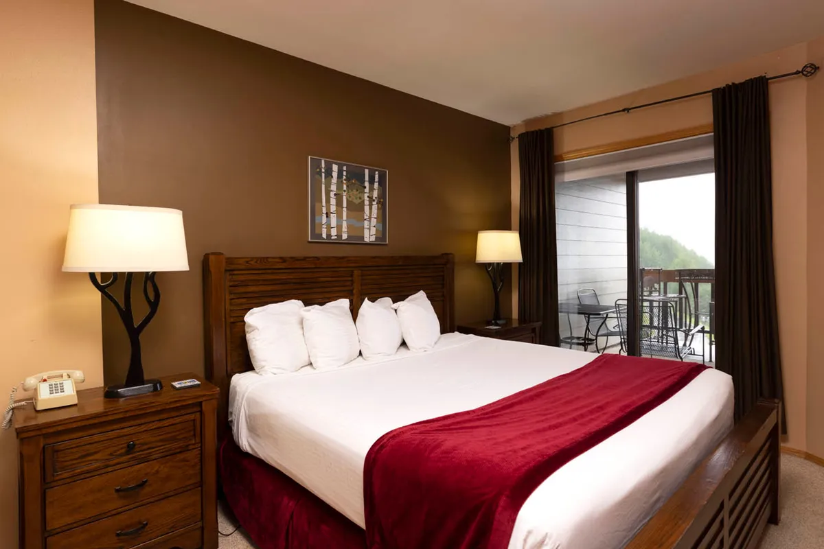 A beautifully designed bedroom featuring a king-sized bed with crisp white linens and a burgundy throw. The scene is softly illuminated by lamps on both sides of the bed, with tasteful artwork above the headboard. Glass doors open onto a balcony where two chairs invite morning coffee or evening relaxation, with a tranquil view beyond.